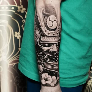 Black and grey Japanese forearm tattoo of a samurai mask with cherry blossoms